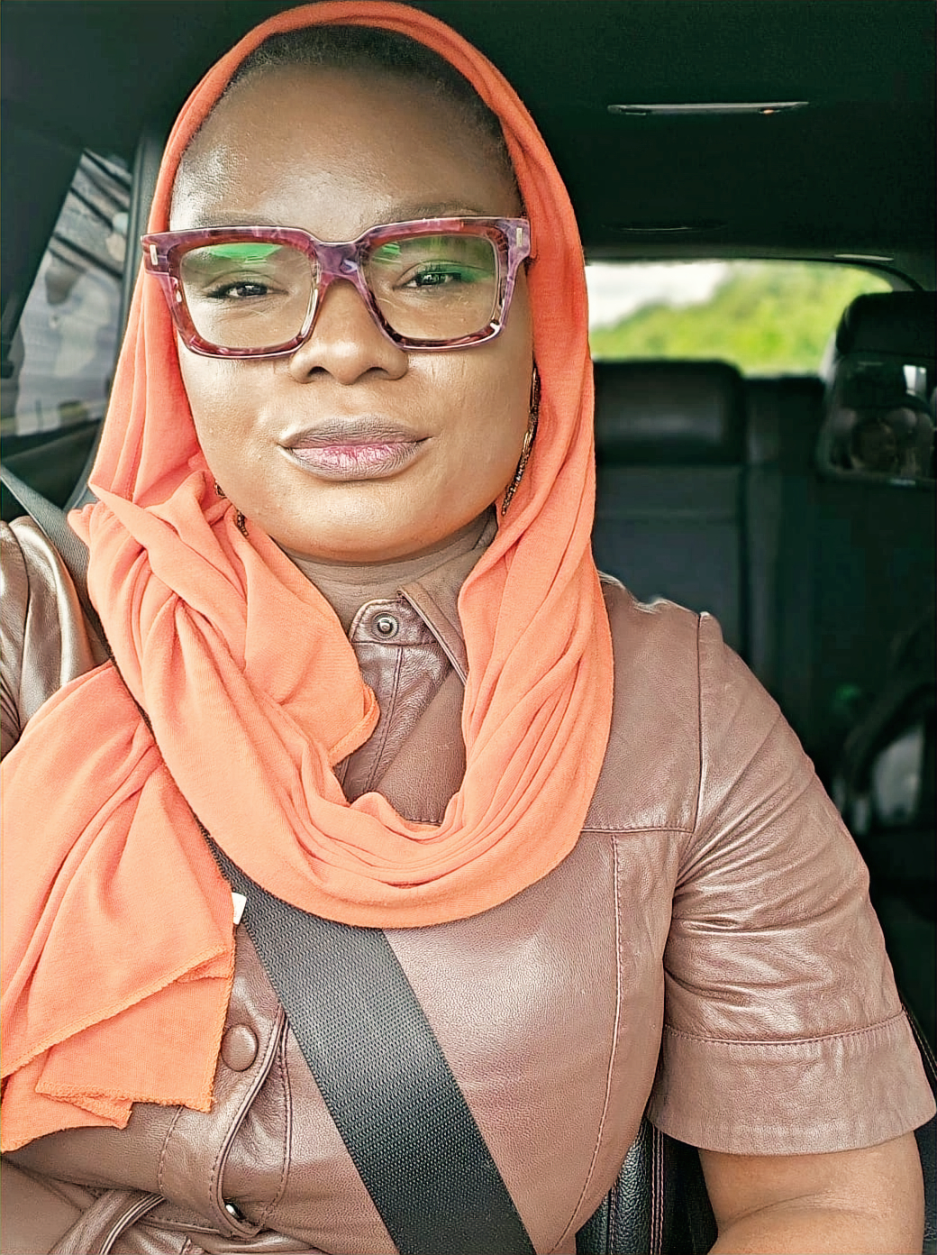 Woman in glasses and orange headscarf in a car, wearing a brown leather top and seatbelt. Calm expression, blurred green scenery outside.